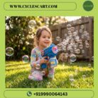 Young girl smiling and sitting on a green lawn playing with the electric blue bubble blaster gun surrounded by bubbles | Cyclescart